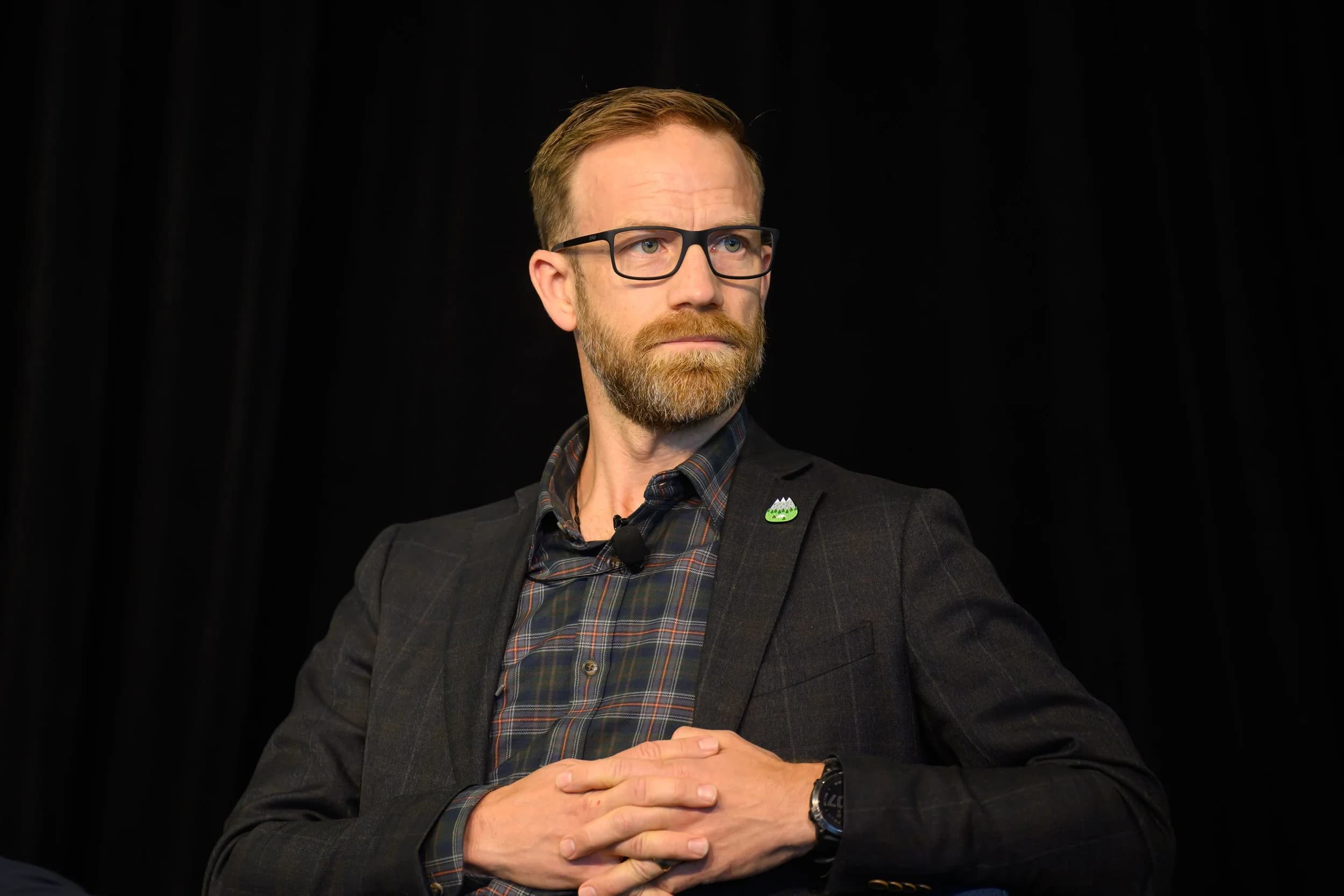 Man with glasses, beard, and short hair wearing a dark blazer and plaid shirt sitting with hands clasped. Black background.