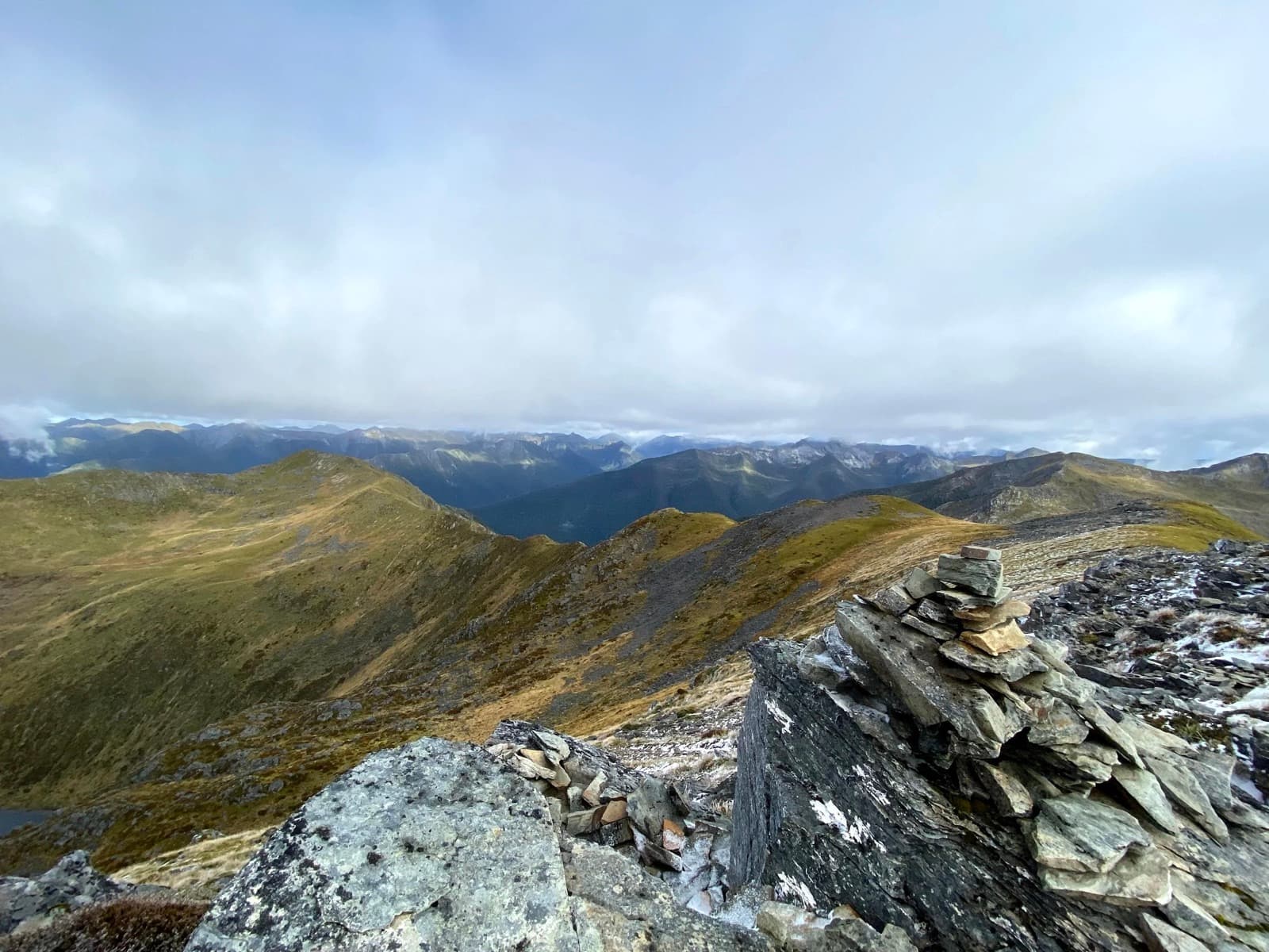 Mountain landscape with grassy ridges and distant peaks under cloudy sky, with a cairn of stacked stones in the foreground.