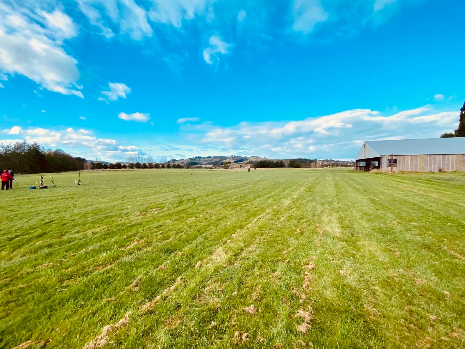 Wide green field with people on the left, a large barn on the right, blue sky with clouds, and distant hills.