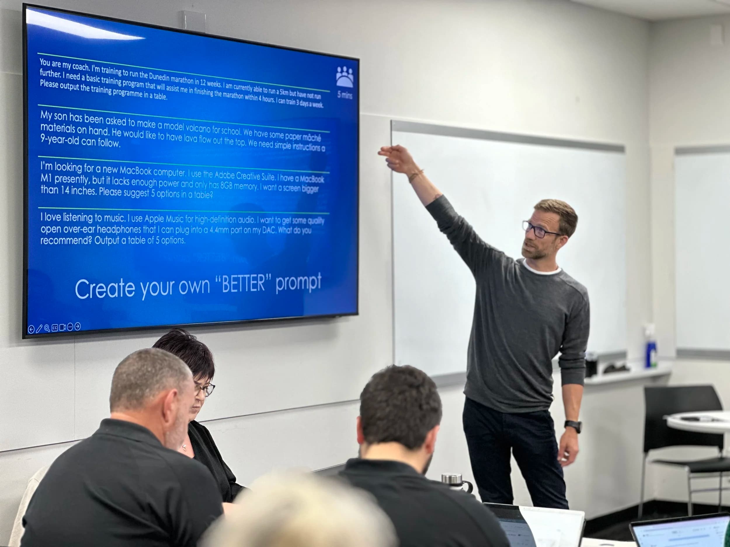 Ben Walker facilitating a hands-on AI workshop, pointing at a screen of real prompts with attendees working on laptops.